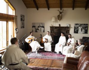 Six Dominicans with instruments sit in a room with a brick fireplace, wood ceiling and musical instruments and recording equipment. Light shines through a large wooded window on the left. 